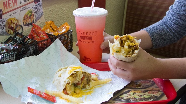 Person holding half of a Del Taco breakfast burrito and a soda