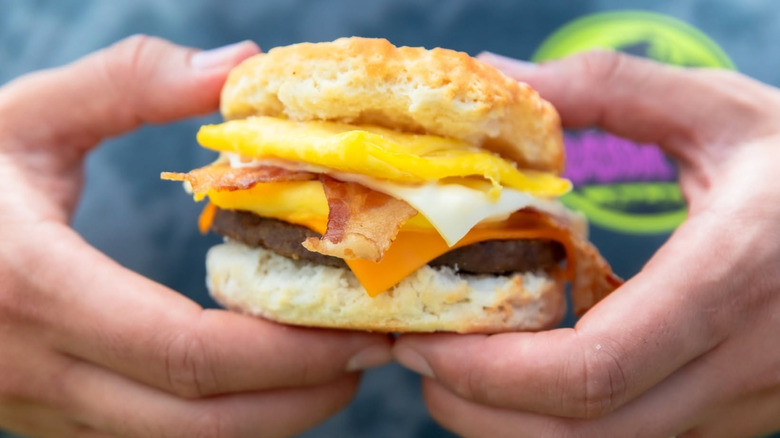 Hands holding a Carl's Jr breakfast biscuit with cheese, egg, bacon, and beef patty