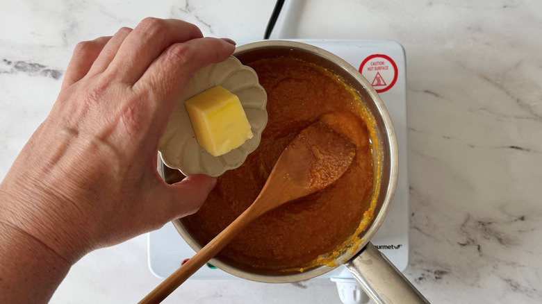 Hand holding small bowl of butter over sauce in pan