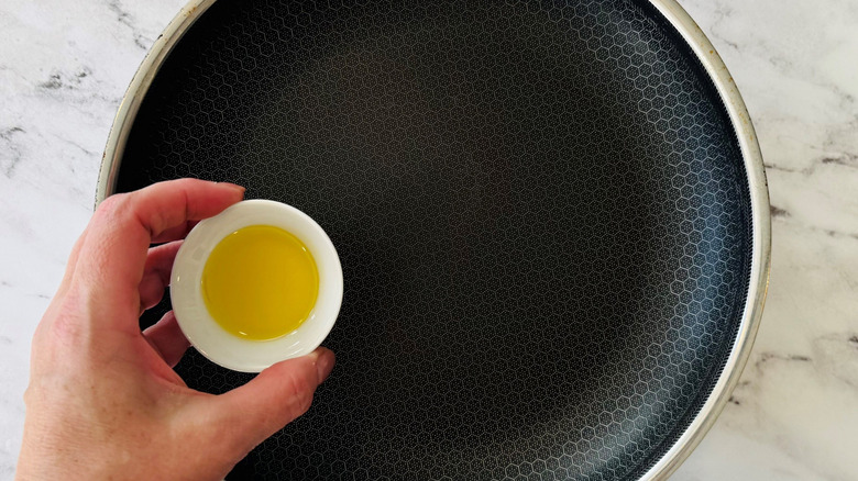 Hand holding small bowl of olive oil over pan