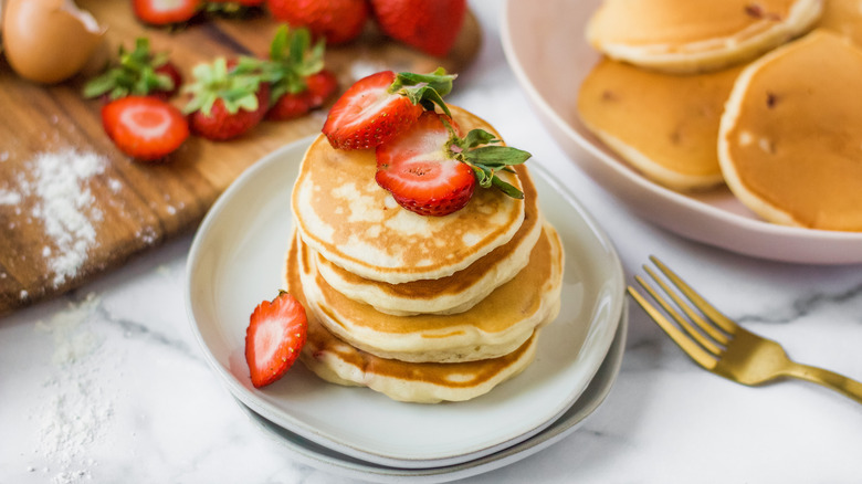 A plate of strawberry pancakes topped with strawberries next to a cutting board with strawberries and a plate with more pancakes