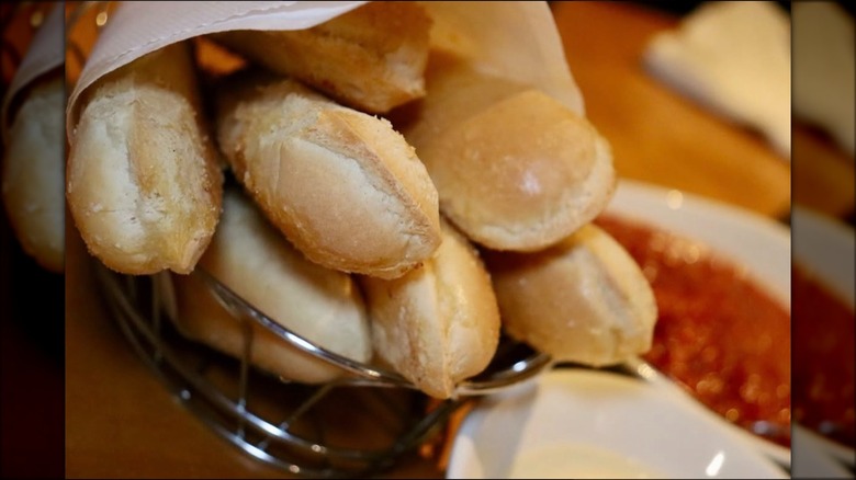A bunch of breadsticks wrapped in a napkin inside a wire basket sitting next to bowls of sauce.