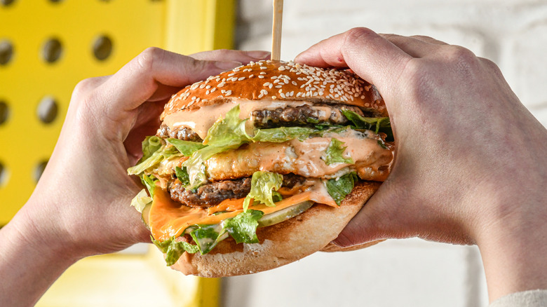 Person holding a massive burger with lots of toppings and sauce with both hands.