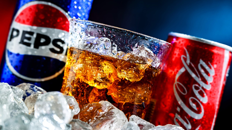 Cola in glass, can of Pepsi blurred on left, can of Coke blurred on right