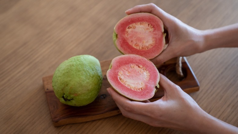 Someone holding guava halves with a whole guava on cutting board.