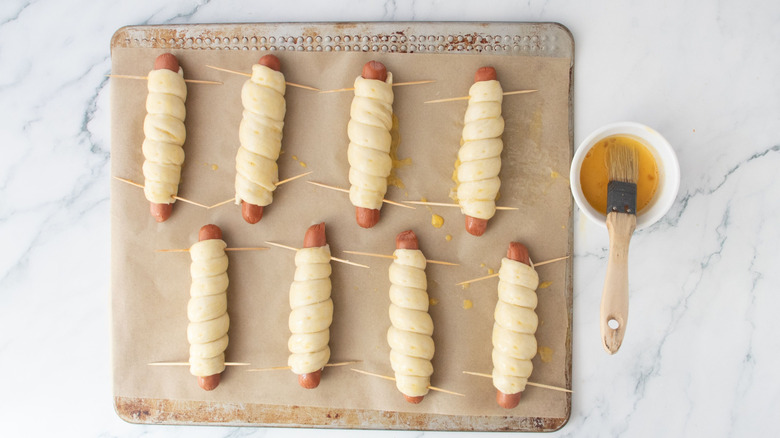 Dough-wrapped hot dogs on baking sheet next to bowl of egg wash