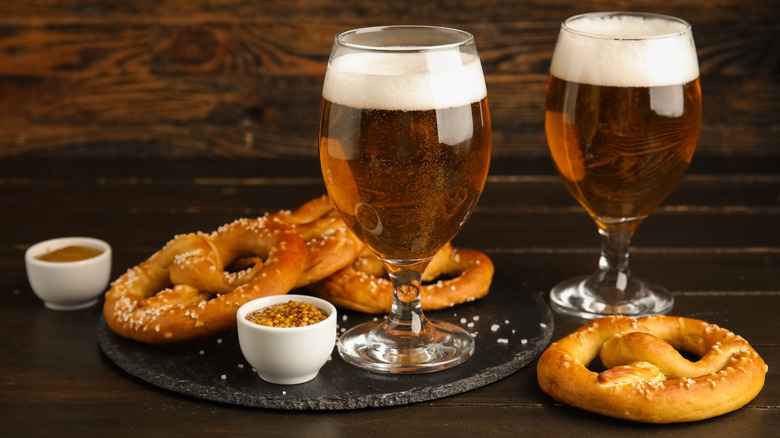 Two German beers in pint glasses with pretzel and mustard.