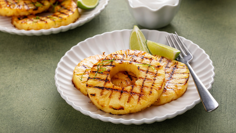 Rings of charred pineapple sit on a plate with lime slices and a fork. A bowl of cream sits in background.