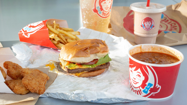 Wendy's burger, chili, fries, chicken nuggets, Frosty, and soda on table.