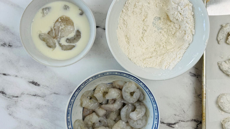 Raw shrimp in bowl next to bowls of flour and buttermilk