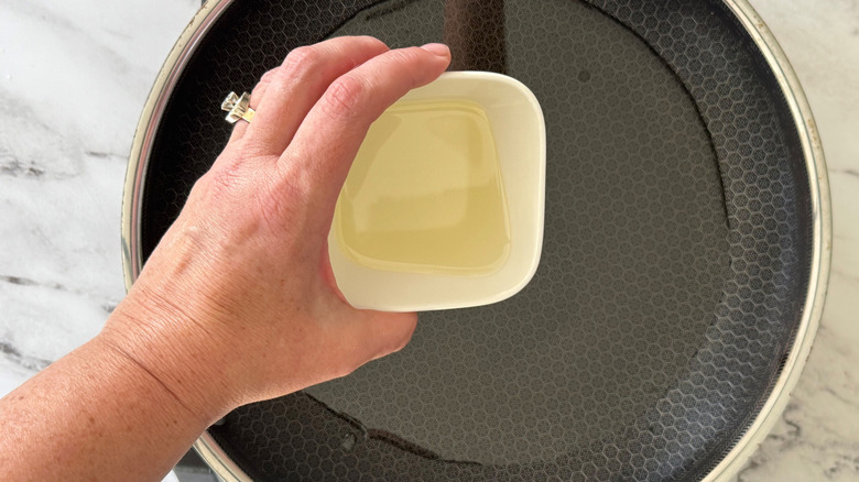 Hand pouring small bowl of oil into pan