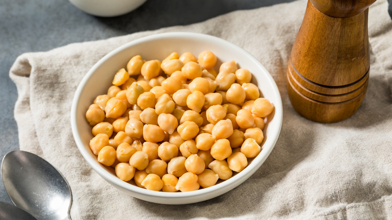 Chickpeas in white bowl on towel next to spoon and wooden grinder