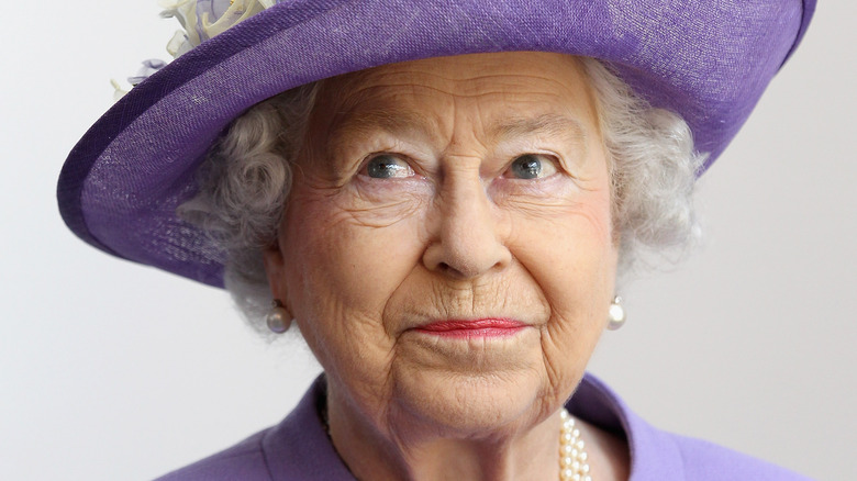  Królowa Elżbieta patrząca w górę i z boku w fioletowym kapeluszu i perłowych kolczykach
