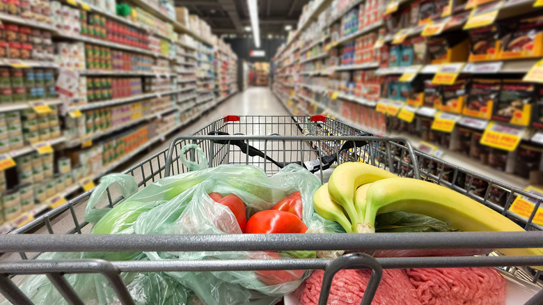A shopping cart holding produce and meat in a grocery store aisle
