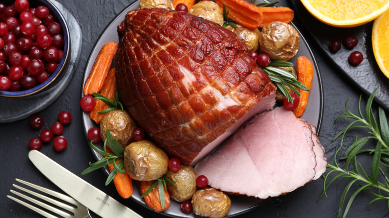 Whole baked ham on a serving tray surrounded by roasted vegetables, fruits, and herbs