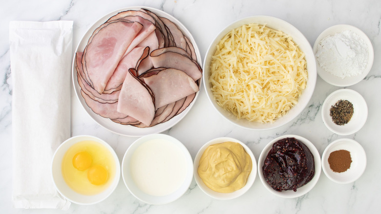 Ham, cheese, mustard, two cracked eggs, jam, and other ingredients for Monte Cristo pinwheels laid out on marble surface