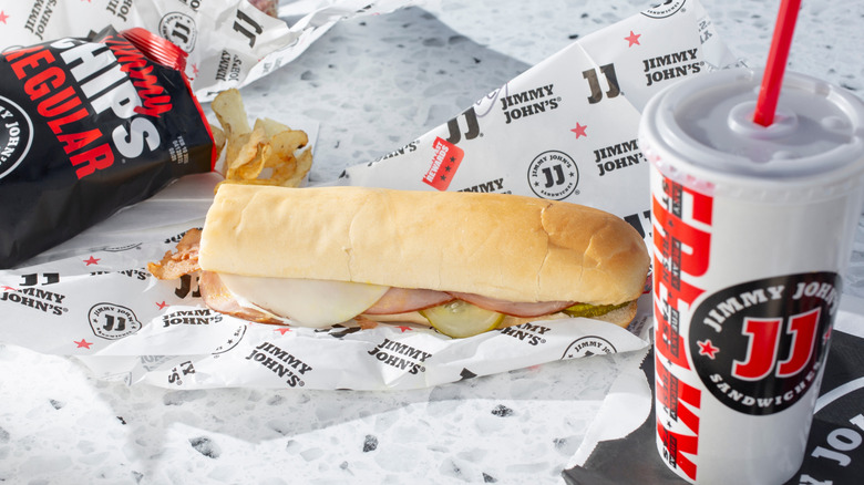 A Jimmy John's sub sandwich, an opened bag of Jimmy's regular chips, and a drink, all on a table