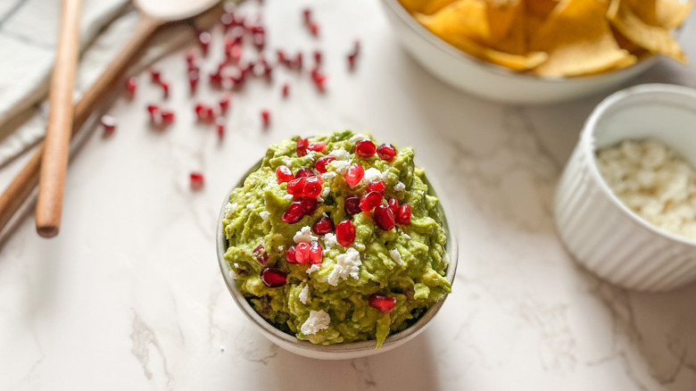 bowl of bright green guacamole topped with juicy red pomegranate seeds near a bowl of tortilla chips, wooden spoons and pomegranate seeds on a counter