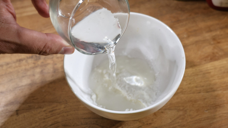 hand pouring water from a small glass into a small white bowl with cornstarch in it