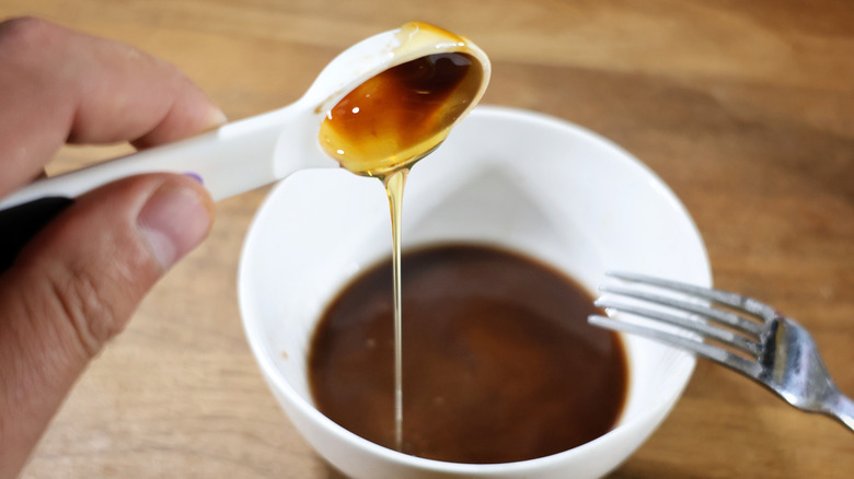 Measuring spoon pouring thin stream of honey into a bowl of dark sauce