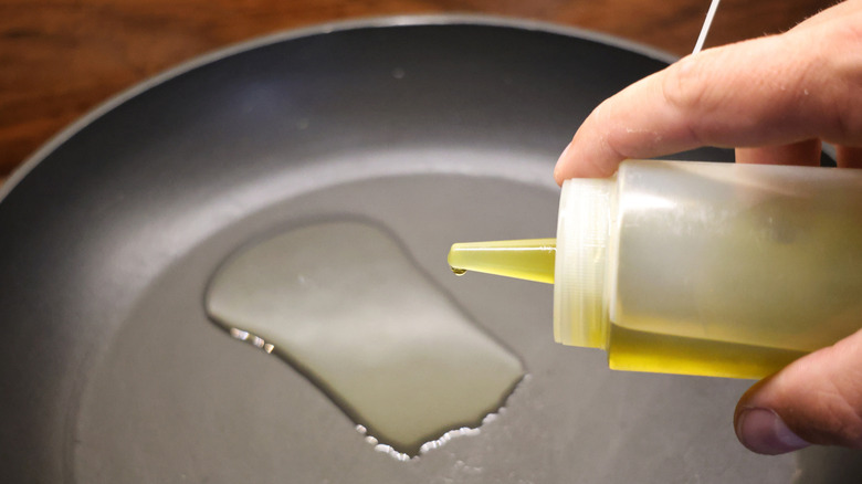 hand holding a squeeze bottle of oil over a skillet with a puddle of oil in it