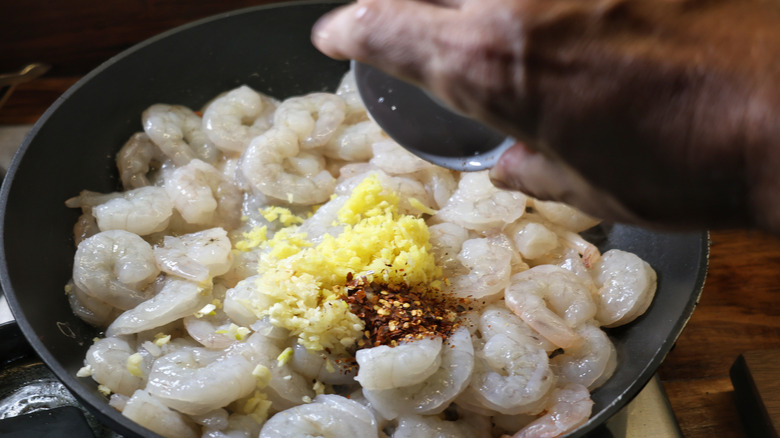 hand dumping a small dish of crushed red pepper into a skillet of shrimp with ginger and garlic