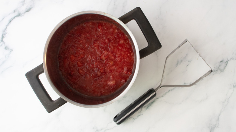 Mashed strawberries in pot next to potato masher
