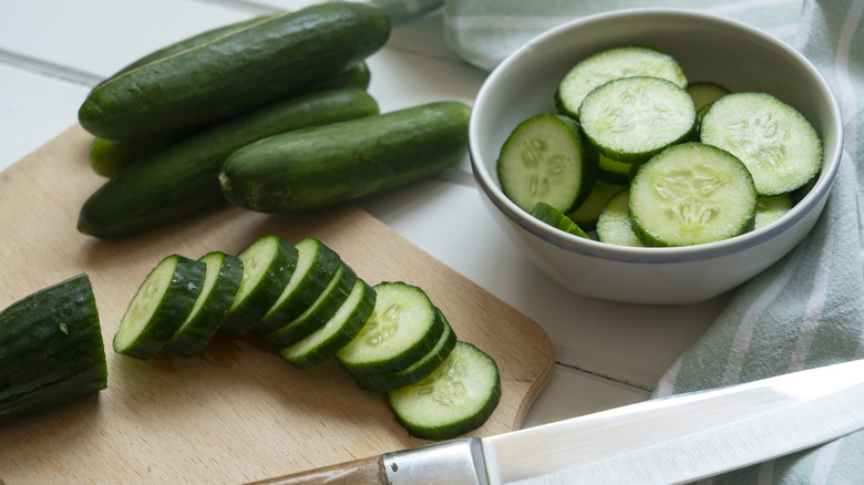 Whole and sliced green cucumbers on a wooden cutting board next to a knife