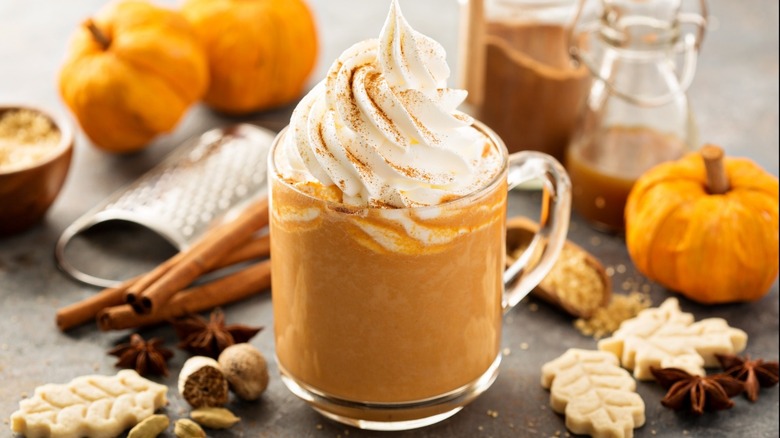 pumpkin spice latte with whipped cream next to pumpkins, cinnamon, nutmeg, spices, and leaf cookies