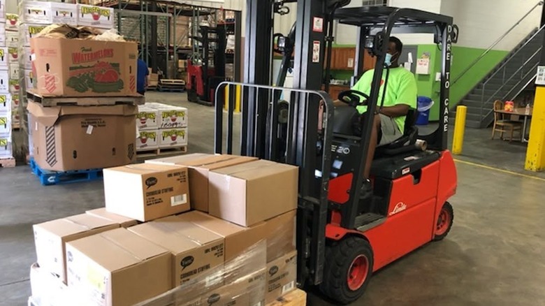 A worker in a lime green shirt drives a red forklift in a food donation warehouse with boxes