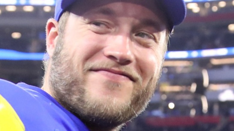 Matthew Stafford smirking in a football stadium wearing a cap