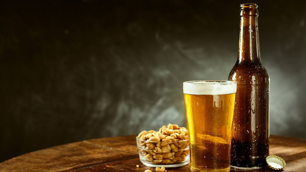 Brown beer bottle, pint glass of beer, glass bowl of nuts and bottle cap on wooden surface