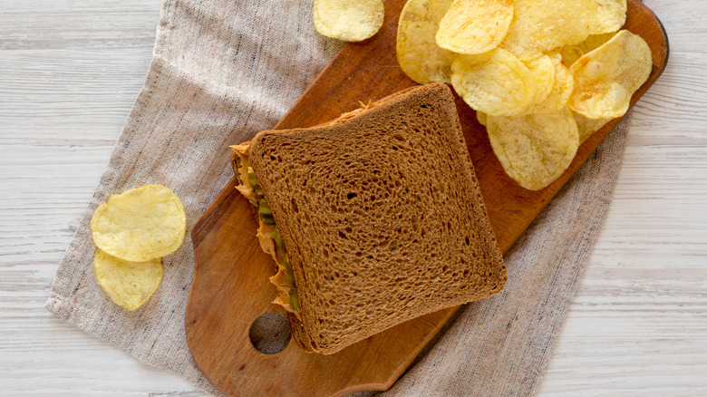 Peanut butter and pickle sandwich with potato chips on wooden board and towel atop gray table