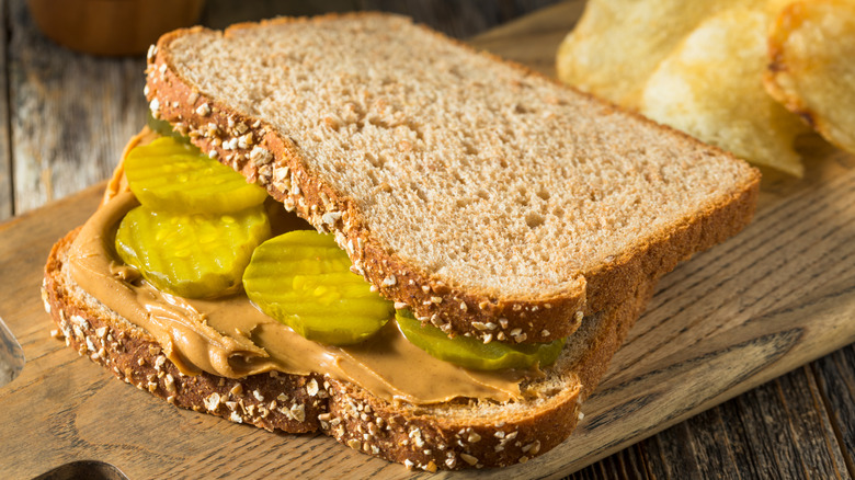 A wheat bread peanut butter and pickle sandwich with chips on a wooden cutting board atop table