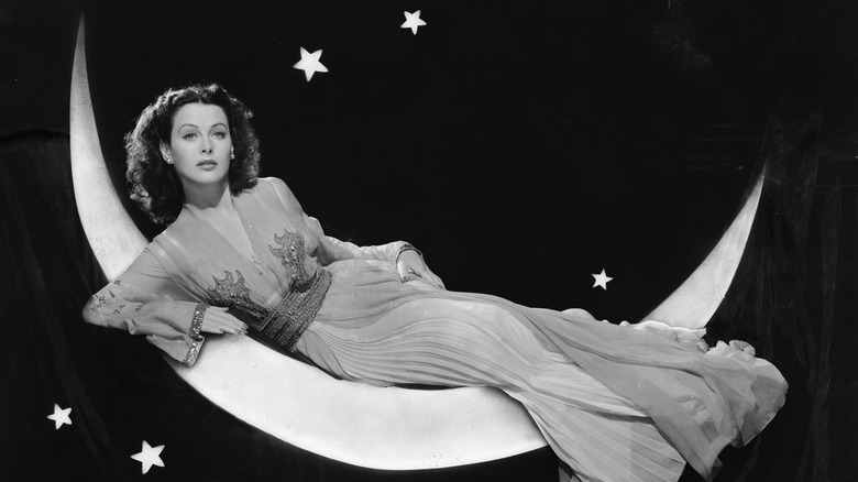 Actress Hedy Lamarr in a long flowing dress, lounging on a prop crescent moon.