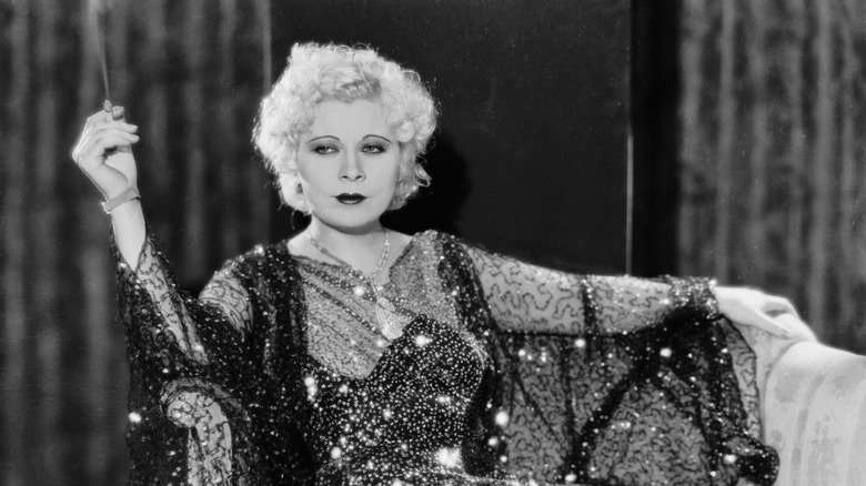 Actress Mae West in a sparkly dress, holding a smoking cigarette, lounging.