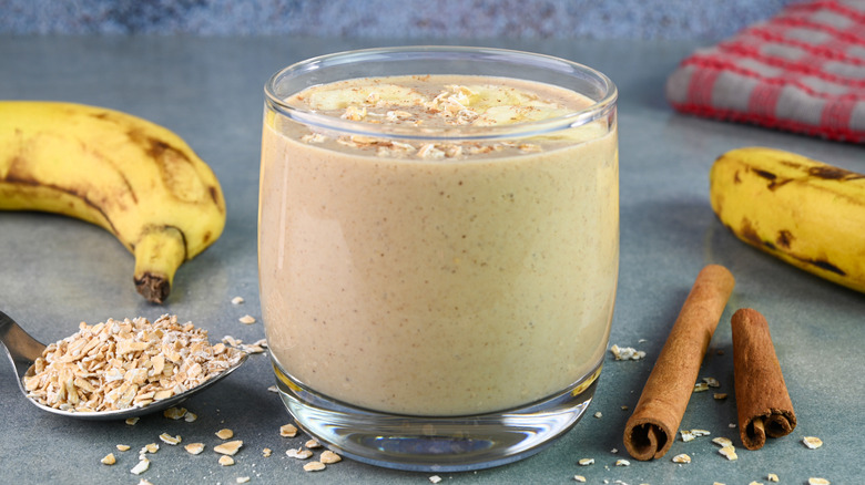 A glass containing a banana oatmeal smoothie with fresh oats on top surrounded by ingredients.