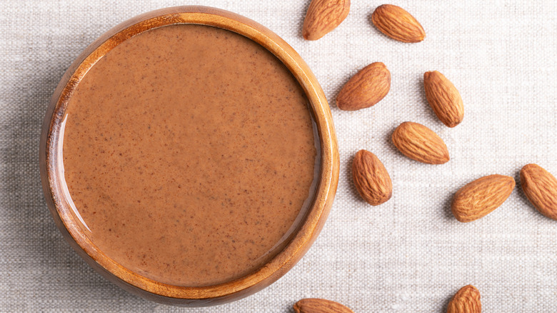 A jar of almond butter next to a few scattered almonds on a white tablecloth
