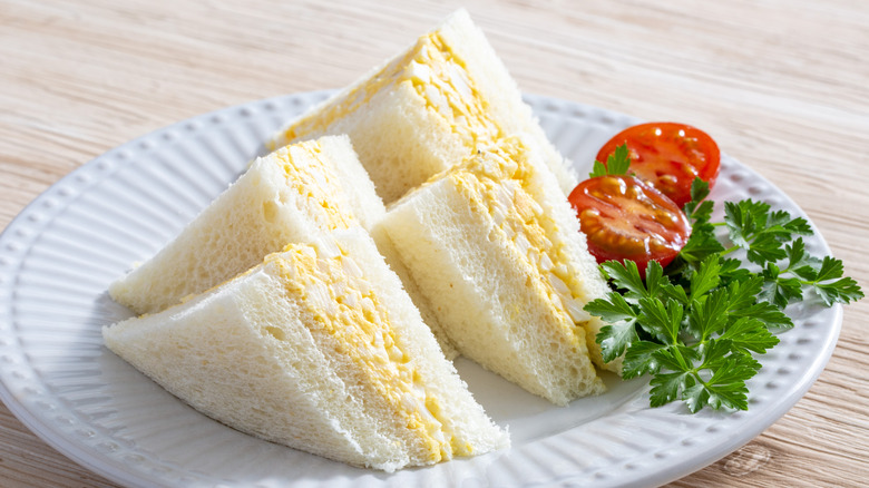 Egg salad sandwiches on white bread on a plate surrounded by sliced tomatoes and a sprig of parsley