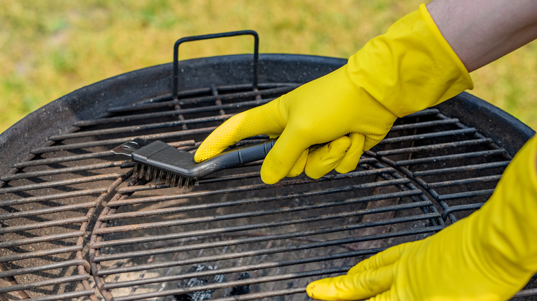 Hands clad in yellow gloves use a brush to clean a grill.