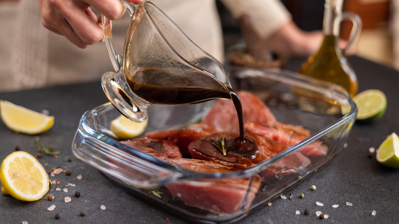 A hand pours marinade from a gravy boat onto fish in a glass pan.