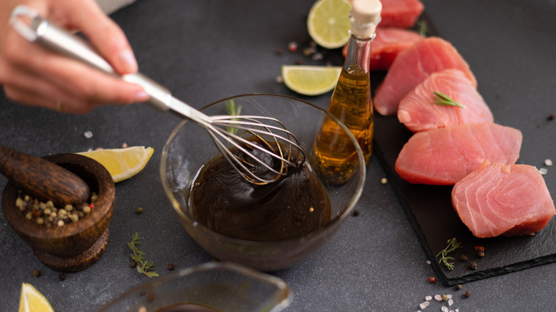 Hand whisking marinade in bowl. Oil, mortar & pestle of peppercorns, lemon slices, and fish surround it.