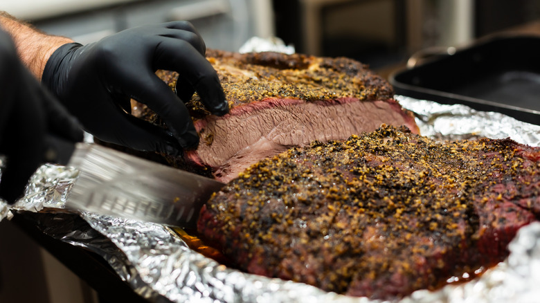 Brisket with herb crust in aluminum foil being sliced by chef's knife