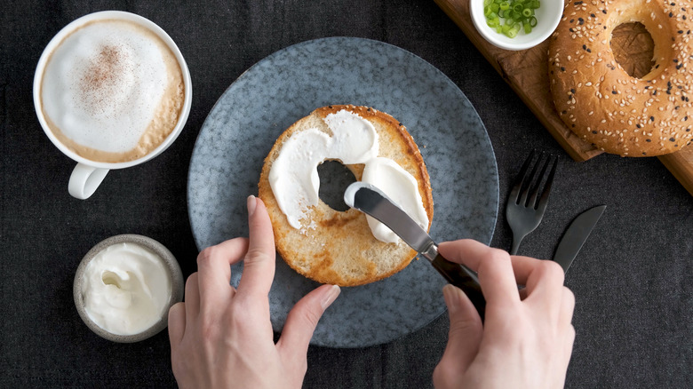 toasted bagel on a plate being spread with cream cheese next to cup of coffee
