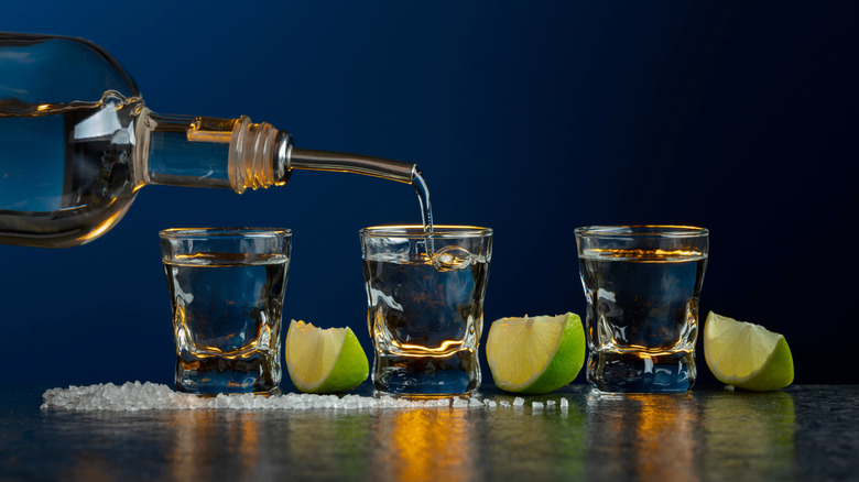 Tequila bottle pouring shots on gray surface with salt and lime wedges against blue background