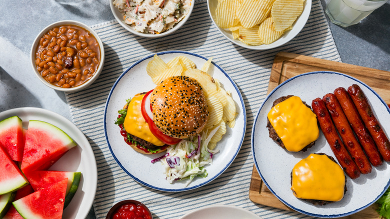 Overhead view of burgers, hot dogs, and cookout sides on picnic table.