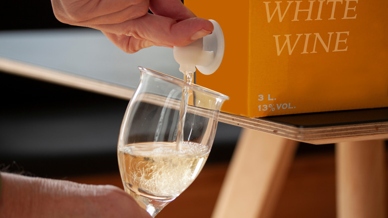 Hands pouring white wine from box on table into glass