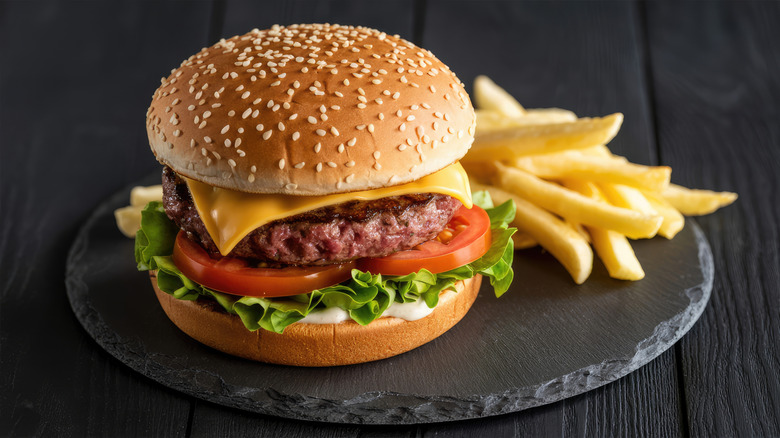 Cheeseburger with tomatoes, lettuce, mayo, and poppy seed bun with fries on black platter and table.