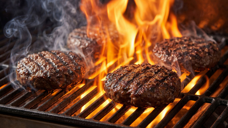 Four burger patties smoking on flaming grill