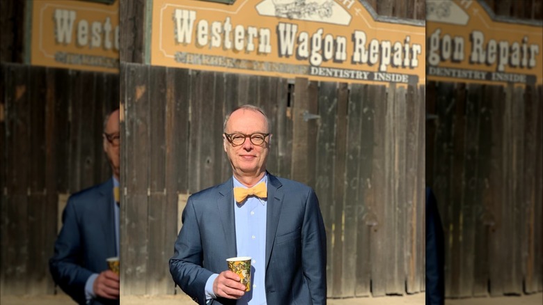 Christopher Kimball holding a drink and smiling in front of a Western Wagon Repair sign